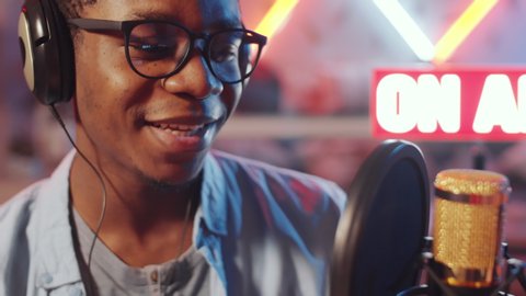 Chest up shot of joyous African American male radio presenter smiling and talking into microphone while broadcasting in studio with lighted on-air sign in the background