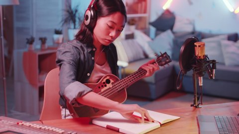 Young Asian female musician in headphones sitting at desk in home recording studio with neon light, playing the guitar and writing lyrics in notepad while creating new song