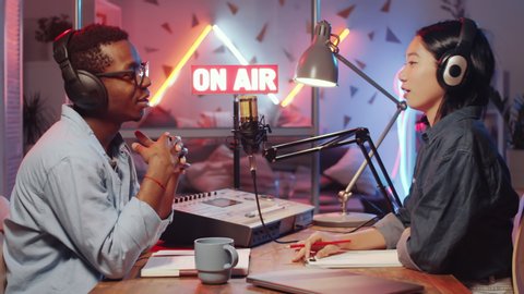 Young Asian woman and Afro-American man sitting together at desk in recording studio with neon light and on-air sign, talking in mic and then taking headphones off and smiling at camera