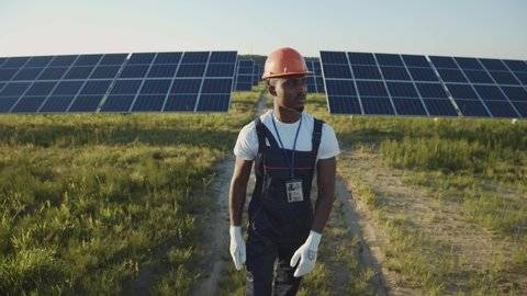 Industrial black man engineer in uniform walking through solar panel field for examination. Ecological construction. Solar power station. Green energy concept.