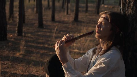 Close-up Man with Long Hair Plays Flute under Pine Trees. On Background Woman with Wreath and Basket Walks in Forest Nature. Slow motion 60 FPS