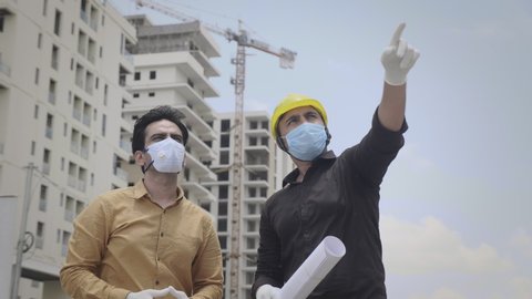 Two young male/ men engineers wearing face protective masks and hand gloves are communicating on a under construction site amid corona virus/ COVID 19 epidemic or pandemic