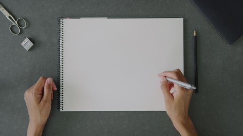 Top view of a woman pretending to write with a pen on a blank a4 sketch book, placed on an office desk with office utensils and notebook.