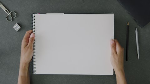 Top view shot of a woman’s perspective holding a blank a4 sketch book, above an office desk with office utensils and notebook.