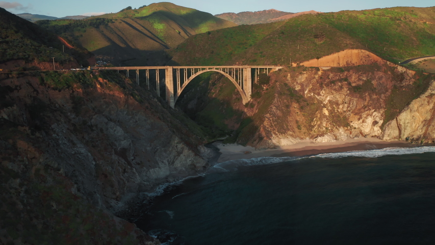 Sunset outdoor. Drone if flying towards the scenic arch bridge at the pure nature of Big Sur park. Panoramic aerial view over the rocky ocean shore. 4K mountains on motion background. Travel USA shot
