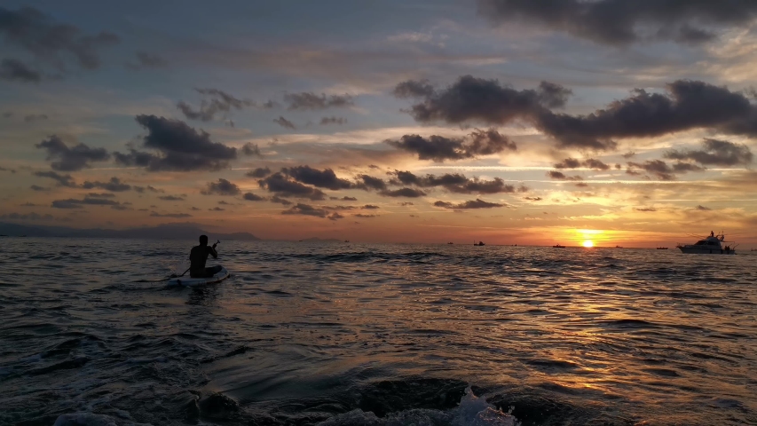 Boy paddling on his paddle while the sun sets.Silhouette of a ship sailing in the background