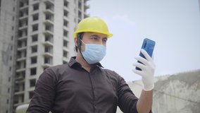 A young male/ man engineer wearing protective face mask and hard hat using mobile phone to talk on an online video call under construction building site amid corona virus/ COVID 19 epidemic/ pandemic