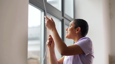 people concept - african american young woman opening window blinds or curtain at home