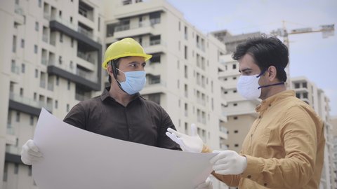 Two young male men engineers wearing face protective mask holding a blueprint of the building and working interacting outside under construction site amid Corona virus COVID 19 epidemic or pandemic