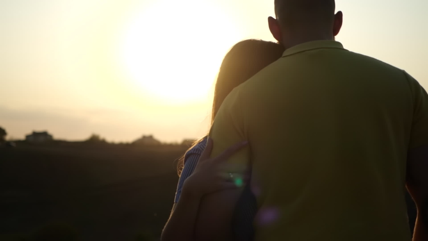 Rear back view of young couple in love, hugging and holding hands against backdrop of sunset in field. Concept of romantic date love in fresh air. Strong guy and girl enjoy beautiful evening in nature Royalty-Free Stock Footage #1056340706