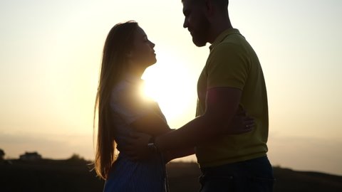 Silhouette of man and woman at sunset. Beautiful girl kisses guy in a deserted field, leaves and rises to hold her hands in slow motion. A romantic date and love on nature of loving happy couple