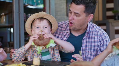 fast food, small attractive boy with straw hat eats mouth watering juicy burger sitting by joyful dad in his arms while relaxing in cafe or restaurant