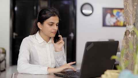 Young woman busy over a phone call while doing work from home on her laptop. Beautiful Indian lady in formal wear, working on her PC while talking on a smartphone - technology concept