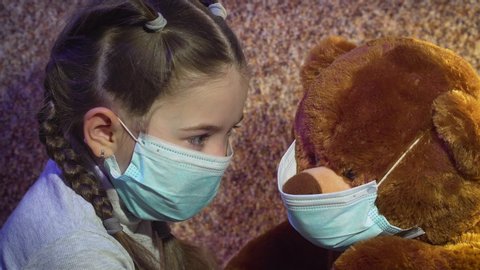 Beautiful girl in medical protective mask stares at her toy bear while staying at home in quarantine. Stop dangerous COVID-19 infection