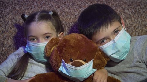 Sad boy and girl in protective face masks sit with their bear toy and look at camera during quarantine. Stop dangerous COVID-19 infection