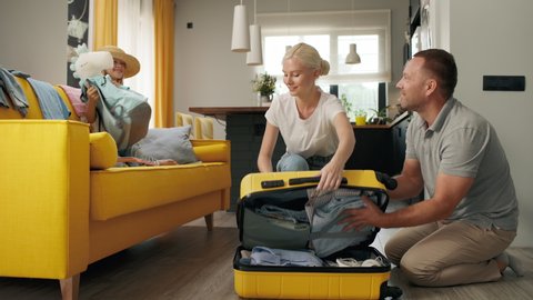 Happy family puts clothes in travel suitcase indoors of home room. Funny smiling girl takes things for summer vacation. Beautiful mom closes luggage before car tour or plane flight. Joy of family love