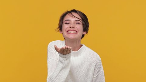 Beautiful cute young woman girl 20s years old in white sweater posing isolated on yellow background in studio. People lifestyle concept. Look camera send blow air kiss showing shape heart with hands