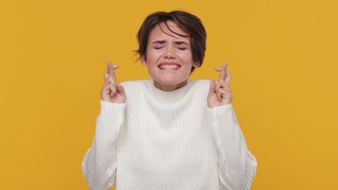 Young woman 20s in white sweater look camera bit lips hands folded in prayer beg about something good keep fingers crossed making wish isolated on yellow background in studio. People lifestyle concept
