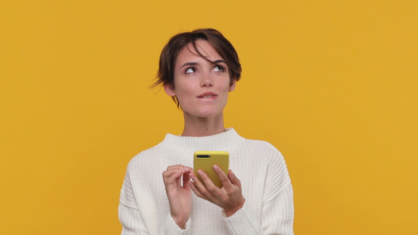 Smiling beautiful young woman girl 20s years old in white sweater posing isolated on yellow background in studio. People sincere emotions lifestyle concept. Look surprised hold using mobile cell phone
