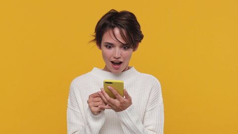 Boring beautiful young woman girl 20s years old in white sweater posing look around get hold using mobile cell phone isolated on yellow background in studio. People sincere emotions lifestyle concept