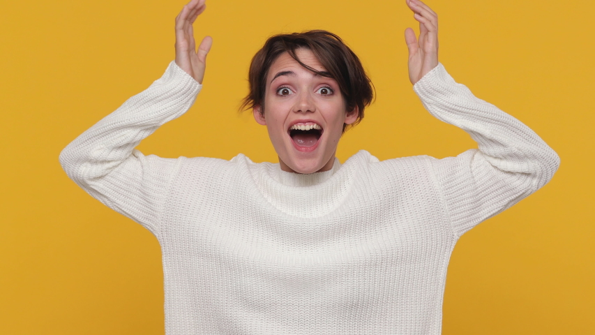 Happy fun beautiful young woman girl 20s in white sweater posing isolated on yellow background in studio. People emotions lifestyle concept. Looking camera shocked surprised say wow put hands on face | Shutterstock HD Video #1056346352