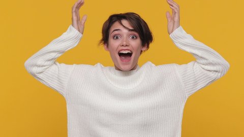 Happy fun beautiful young woman girl 20s in white sweater posing isolated on yellow background in studio. People emotions lifestyle concept. Looking camera shocked surprised say wow put hands on face