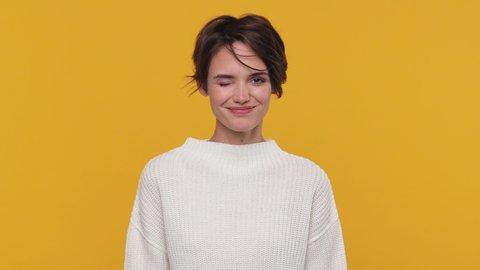 Smiling beautiful young woman girl 20s years old in white sweater posing isolated on yellow background in studio. People sincere emotions lifestyle concept Looking camera charming smile wink blink eye
