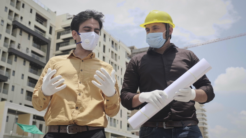 Two young male men engineers wearing face protective mask holding a blueprint of the building and working interacting outside under construction site amid Corona virus COVID 19 epidemic or pandemic | Shutterstock HD Video #1056347963