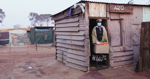 Close-up of an old Black African woman standing in the door way of her tin shack in an informal settlement with a food parcel donated to her by an NGO during Covid-19