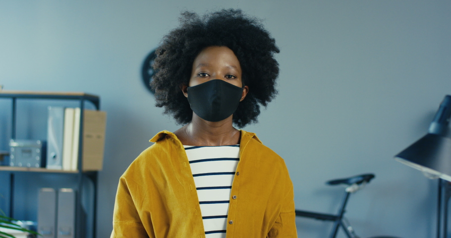 Close up portrait of beautiful cheerful African American girl putting off protective mask at office indoors. Pretty female employee in cabinet smiling to camera. Work in quarantine. Work concept | Shutterstock HD Video #1056348749