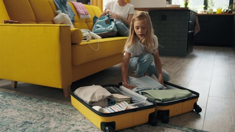 Happy family puts clothes in travel suitcase indoor of new home room. Pretty girl and attractive sister take things for summer holiday at car tour or prepare baggage for relocation. Joy of family love