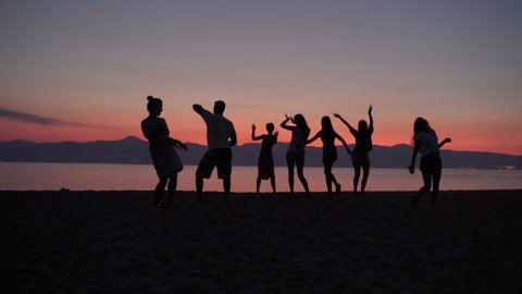 Beautiful sea sunset background, crowd of happy cheerful friends dancing barefoot at the sea beach. Silhouettes of dancing people lifting uptheir hands. Beach party background