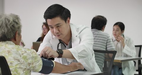 Smiling asian male doctor checking blood pressure test to the elderly female. A group of young adults volunteer nurse doing basic health care clinics for senior patient at local volunteer center. 