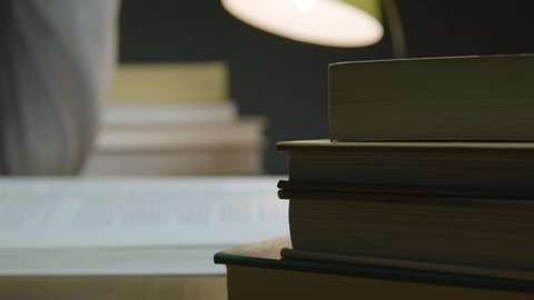 A close up shot of a stack of books in the foreground with a male reading and turning a page of a book in the background, shot in a dark room with a lamp in shot.