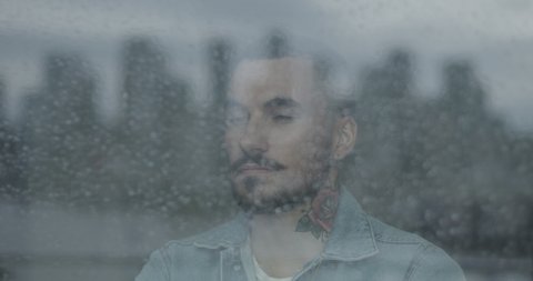Hipster bearded young adult male looking out of window on rainy day at view of city skyline