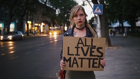 Lady hipster is showing cardboard poster, inscription all lives matter. She posing standing by a road, city with busy evening traffic. Dark cinematic