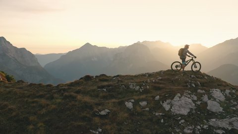 Aerial - Drone flying backwards revealing the adult mountain rider sitting on his bike on top of the mountain peak at sunset