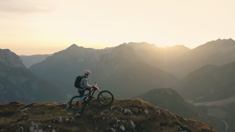 Aerial - Mountain biker with a backpack arriving to the mountain peak enjoying the last sun rays shinning on the majestic mountains. 4K