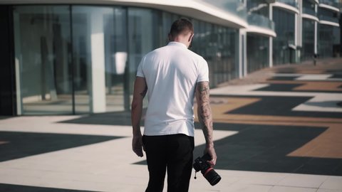 Backside view, tattooed male photographer in white t-shirt walking with camera in hands along street, cityscape on background. High quality FullHD footage