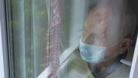 bald pensioner in blue facial mask blurry reflection in plastic window glass behind transparent curtains slow motion closeup