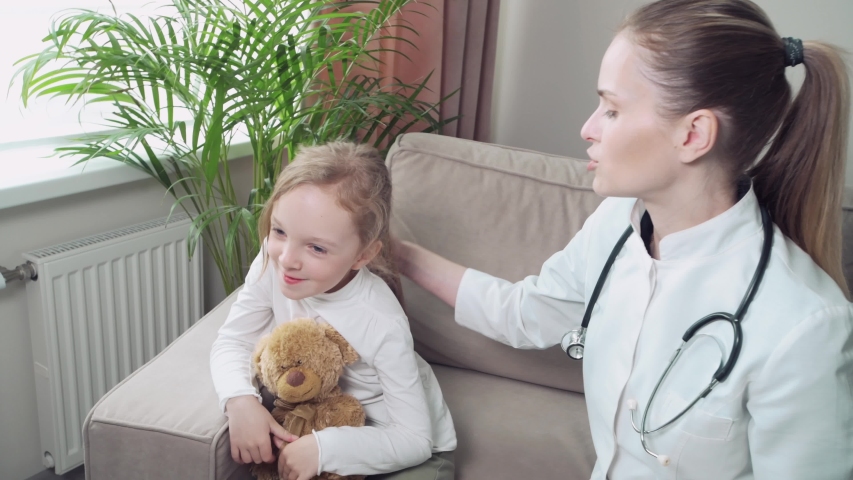 Young beautiful woman doctor examines a sick child at home, sitting on a sofa.