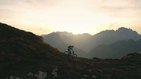 Aerial - Skilled mountain biker cycles down the steep terrain, comes to the mountain edge and enjoys the view of beautiful nature at sunset. 4K
