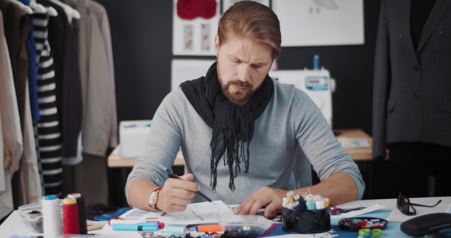 Handsome man sitting at table with sewing equipments, looking aside and thinking about new ideas. Fashion designer working with sketches at modern studio. | Shutterstock HD Video #1056377228