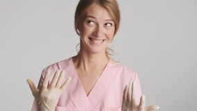 Young cheerful female doctor in medical gloves slyly showing come here gesture on camera over gray background