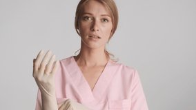 Young beautiful female doctor confidently wearing medical gloves smiling on camera over gray background