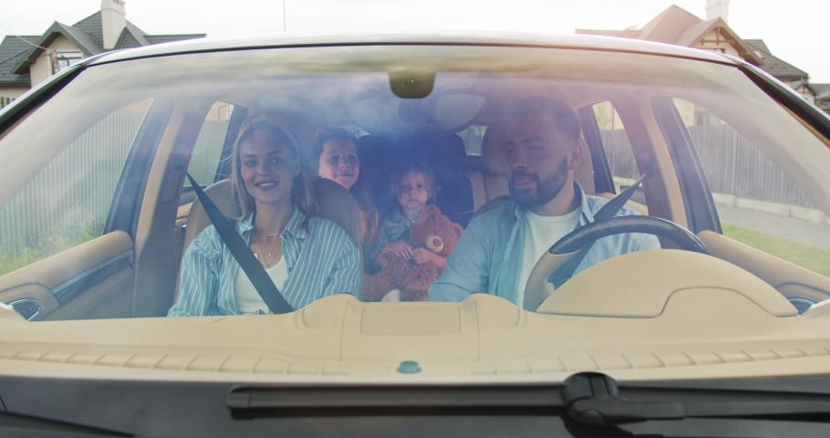 Front view of parents and excited doughters smiling driving car in city. Young family waving to new neighbors after successful home purchase. Concept of lifestyle, happy family, travel together. Royalty-Free Stock Footage #1056379160