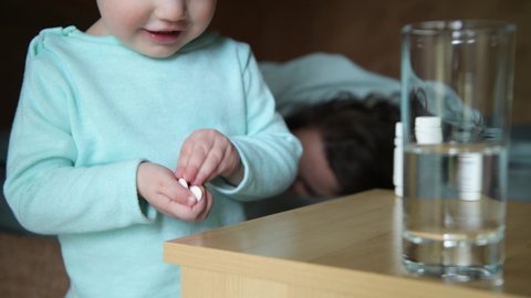 A small child comes up and takes pills from the bedside table on the background of a sleeping mother. The danger of poisoning, the carelessness of parents. Close up