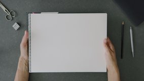 Top view shot of a woman’s hands holding a blank a4 sketch book above an office desk with office utensils and notebook. - Powered by Shutterstock - Get 15% off with code: PIKWIZARD15