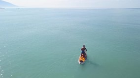 Aerial drone view of a man practicing stand up paddle or SUP in tropical exotic sea. Video. Flying over the turquoise calm sea and a man standing on a sup board on mountains and cloudy sky background.