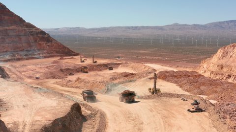 Cinematic aerial view of the red industrial landscape of the open pit with blue sunny sky. Windmill and turbine tower farm is visible down in the desert valley. Large industry business site, USA 4K